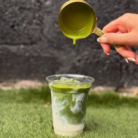 Bright green matcha being poured from a gold jug into an iced milk, set on the grass