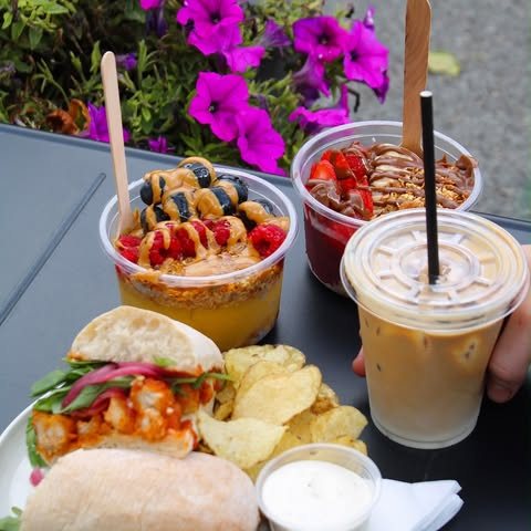 A café table in the sun with two açaí cups, an iced latte, a loaded sandwich and crisps, pink flowers behind
